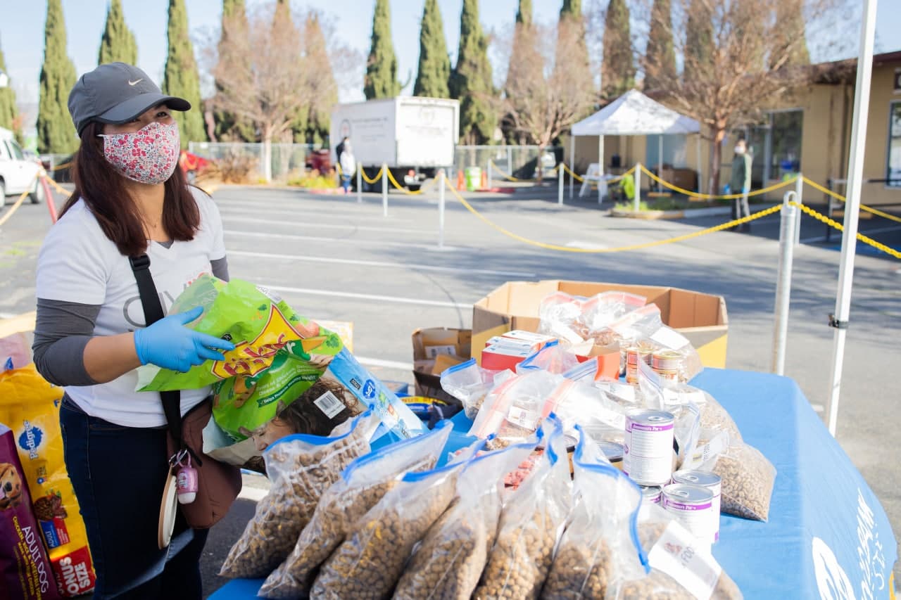 Woman with face mask standing next to table full of dog food and supplies.